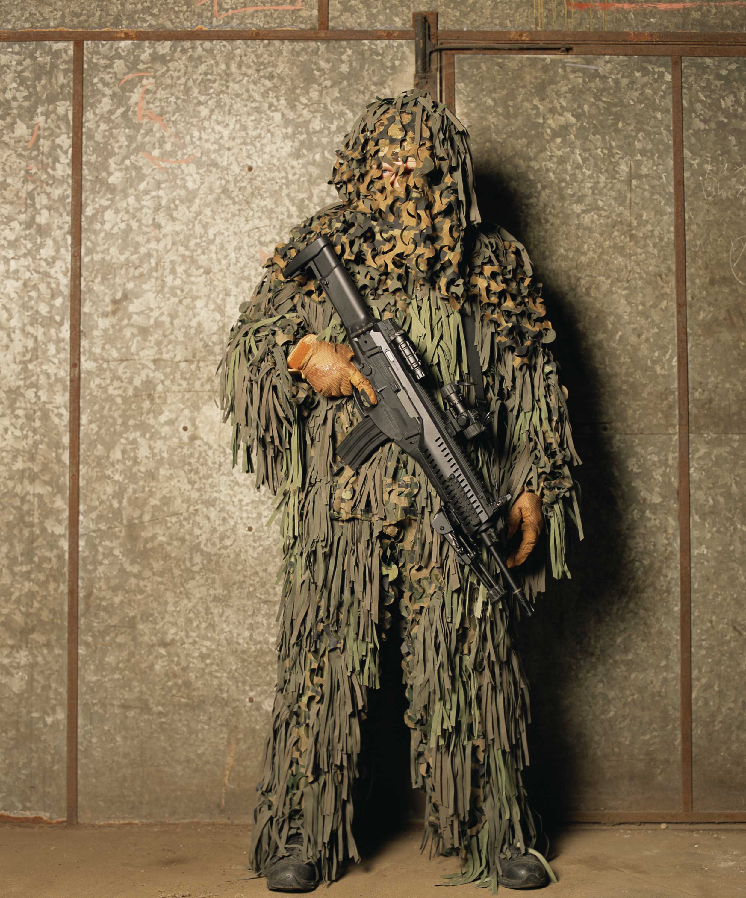 A photograph by the artist Andres Serrano titled 'Man in Camouflage Suit'. It features a person in a green camouflage suit that mimics the texture of a tree. They are holding an assault rifle and stand against a concrete wall.