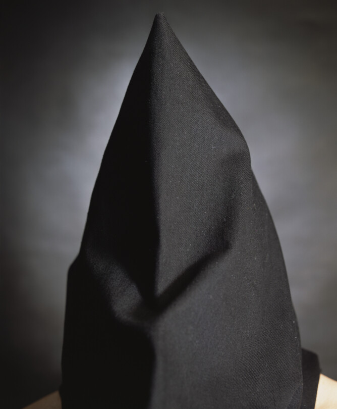 Photo by artist Andres Serrano titled 'Kevin Hannaway, "The Hooded Men"'. It features a black hooded figure with bare shoulders, stood against a grey background.