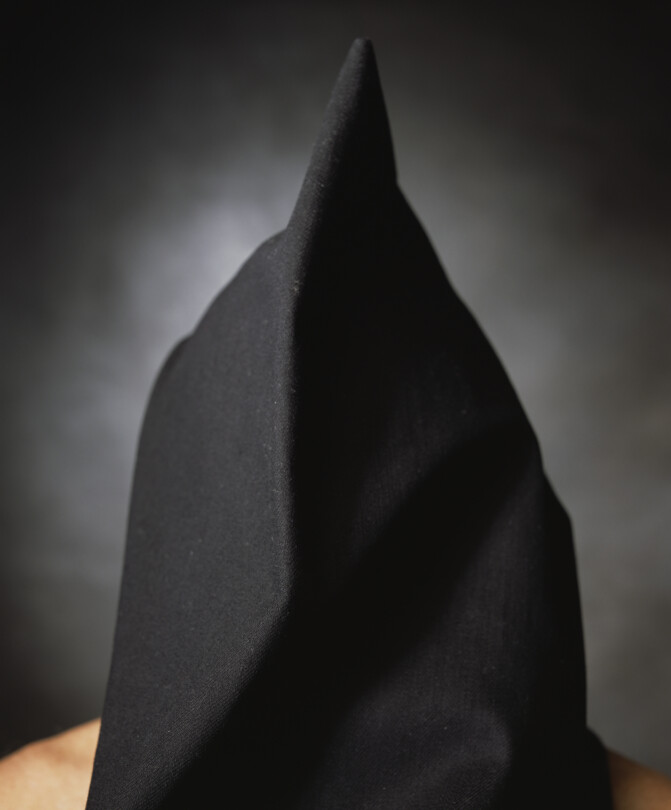 Photo by artist Andres Serrano titled 'Patrick McNally, "The Hooded Men"'. It features a black hooded figure with bare shoulders, stood against a grey background.
