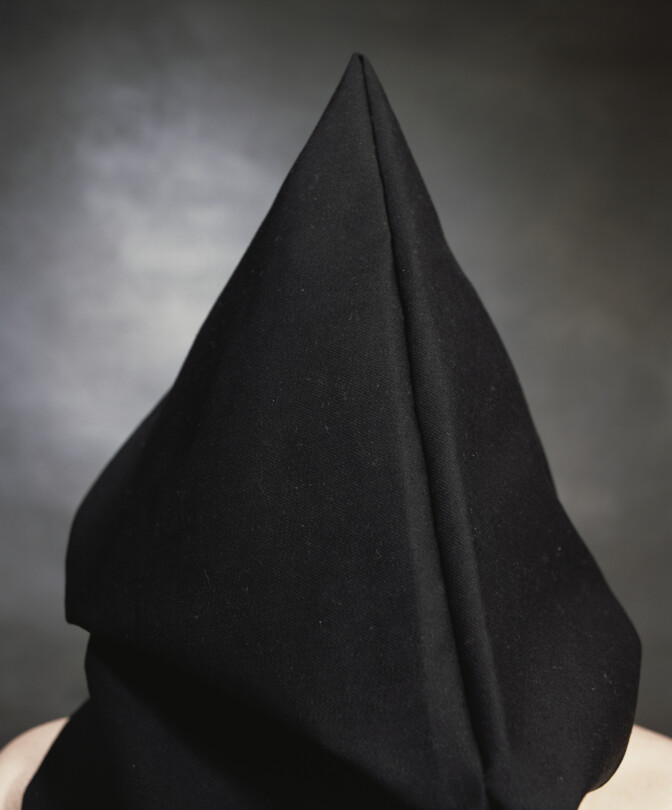 Photo by artist Andres Serrano titled 'Brian Turley, "The Hooded Men"'. It features a black hooded figure with bare shoulders, stood against a grey background.