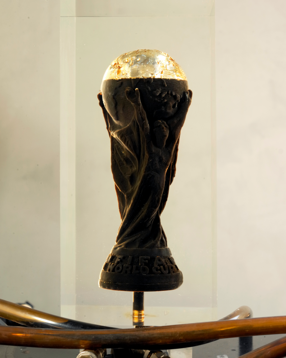 This image depicts Andrei Molodkin's sculpture The Fifa World Cup Filled With Qatari Oil. The artwork is an acrylic block with a mould of the Fifa World Cup, this block is connected to pipes that trail at the bottom of the screen, the cup is nearly full with crude oil.