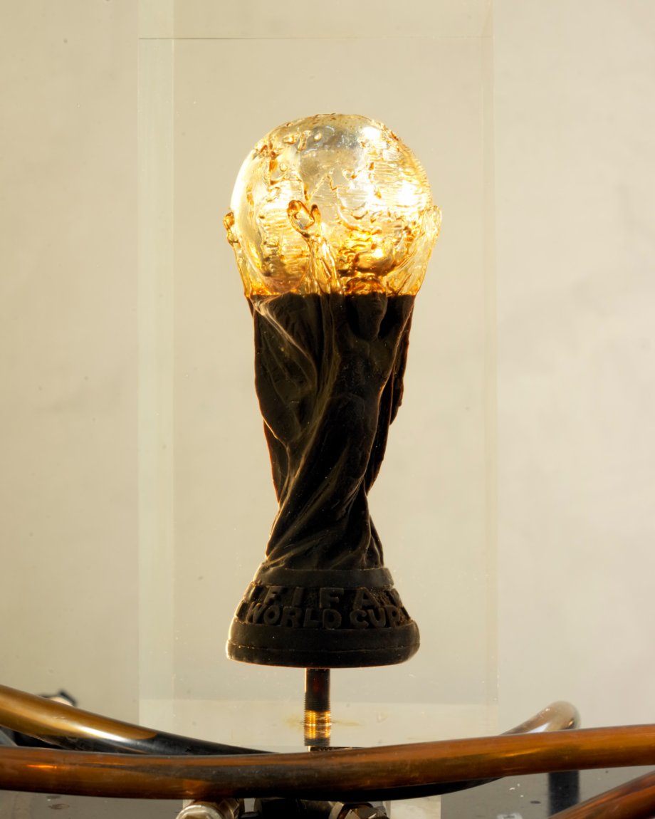 This image depicts Andrei Molodkin's sculpture The Fifa World Cup Filled With Qatari Oil. The artwork is an acrylic block with a mould of the Fifa World Cup, this block is connected to pipes that trail at the bottom of the screen, the cup is two-thirds full with crude oil.