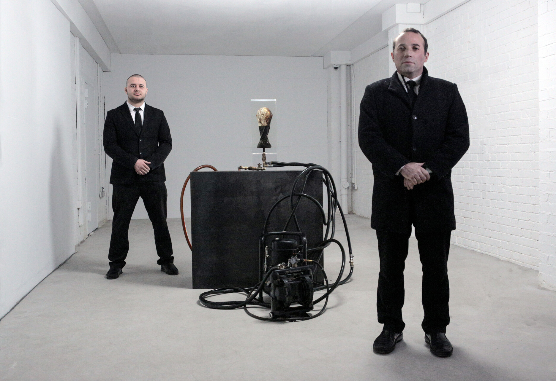 This image depicts Andrei Molodkin's sculpture The Fifa World Cup Filled With Qatari Oil being guarded by two stern security guards. The artwork is an acrylic block with a mould of the Fifa World Cup, this block is connected to a sea of pipes that trail from a central black podium onto the floor.