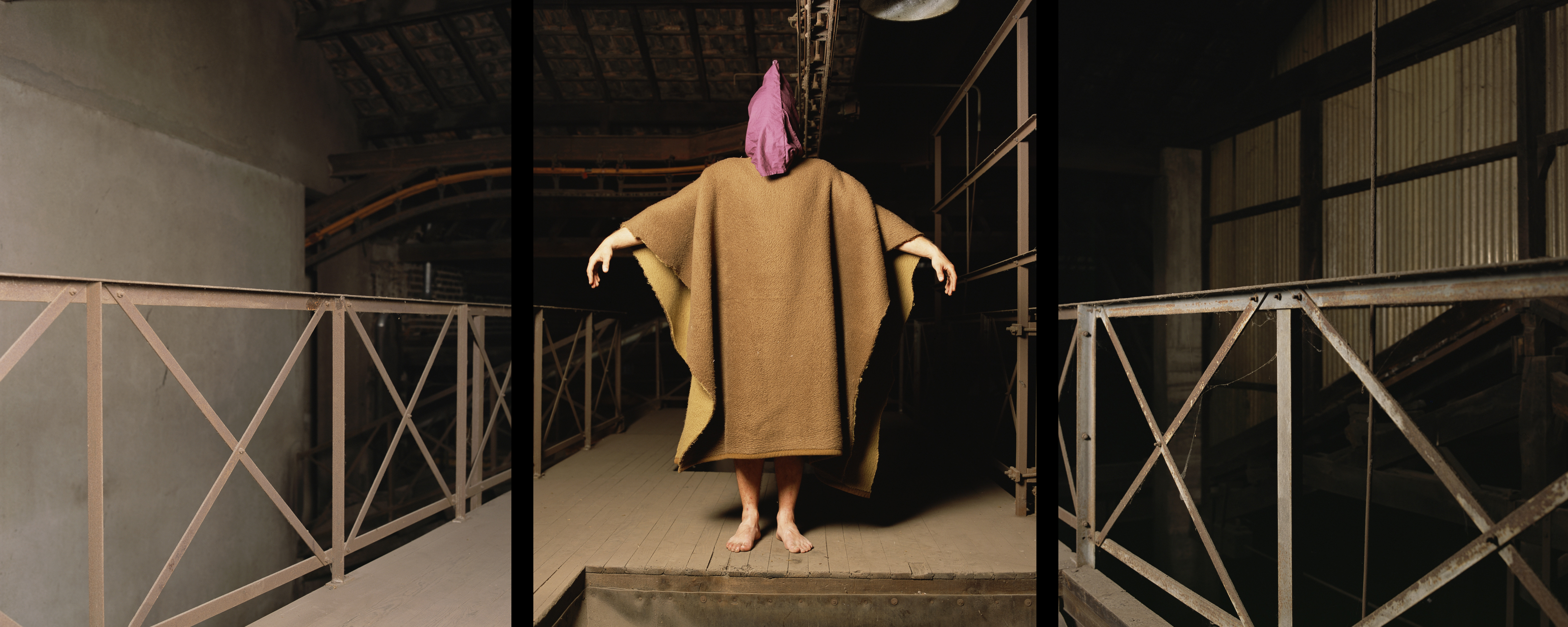 A triptych by the artist Andres Serrano depicting a hooded figure clothed in a rag in the central image. The figure is holding their arms up to form a cross whilst standing in the middle of a raised walkway. The exterior is of a dark factory setting.