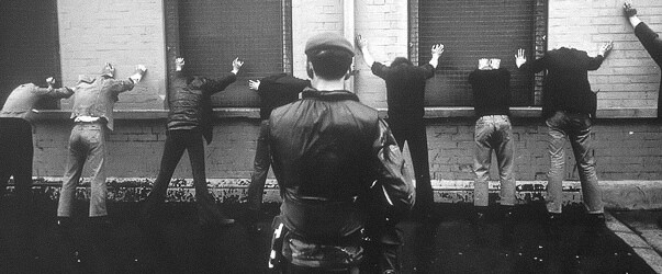 A historical photo of a British soldier in Northern Ireland during 'The Troubles' pushing a group of 7 people against a wall at gun point. These figures are stood with their hands against a brick wall and their legs spread shoulder width apart.