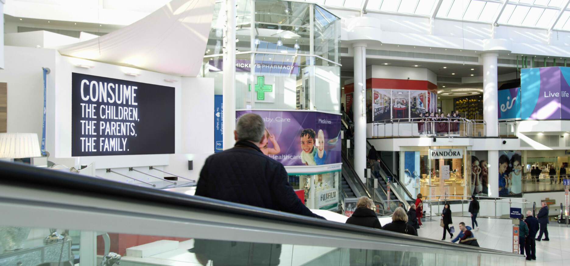 An image from Act II of ORDER, featuring a populated shopping mall, with an advertising board which reads: "Consume the children, the parents, the family."