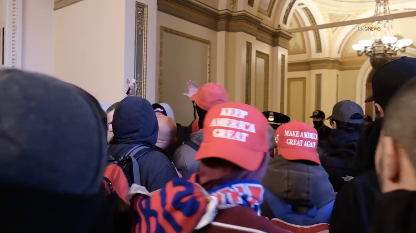 A still from INSURRECTION, a film by Andres Serrano. The still features Trump supporters roaming the corridors of Capitol Hill. A red hat reads: "Make America Great Again".