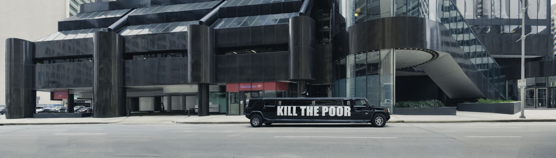 An artwork by Democracia from Act I of ORDER. This is a panoramic photo of a black stretch limo with the words KILL THE POOR in white on the side. The limo is moving through a city street in a financial district.