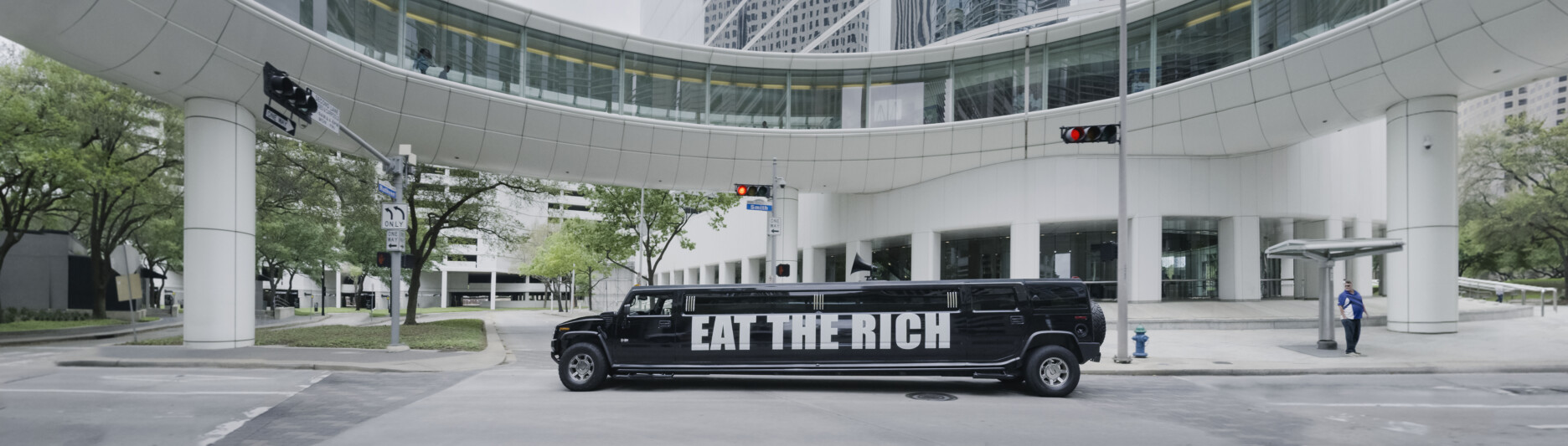 An artwork by Democracia from Act I of ORDER. This is a panoramic photo of a black stretch limo with the words EAT THE RICH in white on the side. The limo is moving through a city street in a financial district.