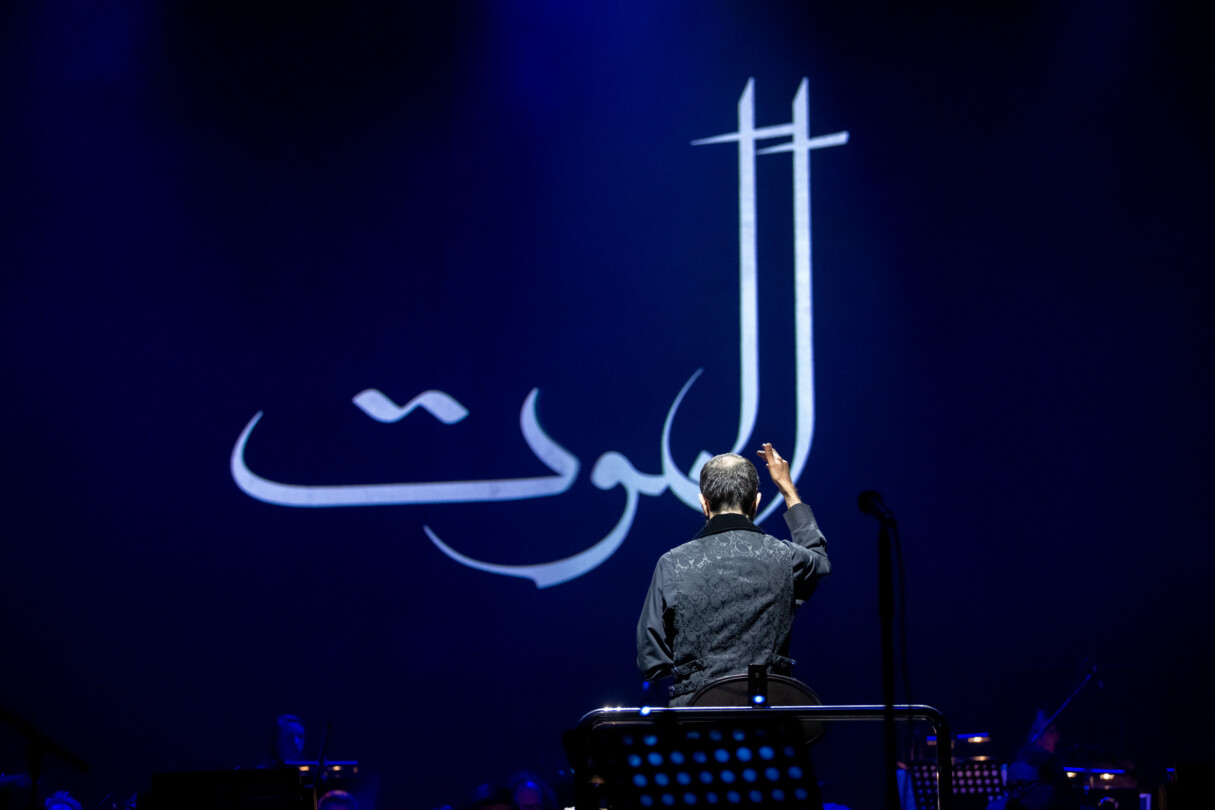 Screenshot from the performance of Alamut by Laibach in Ljubljana, Slovenia. This image depicts the conductor in front of a large Arabic lettering which reads 'ALAMUT'.