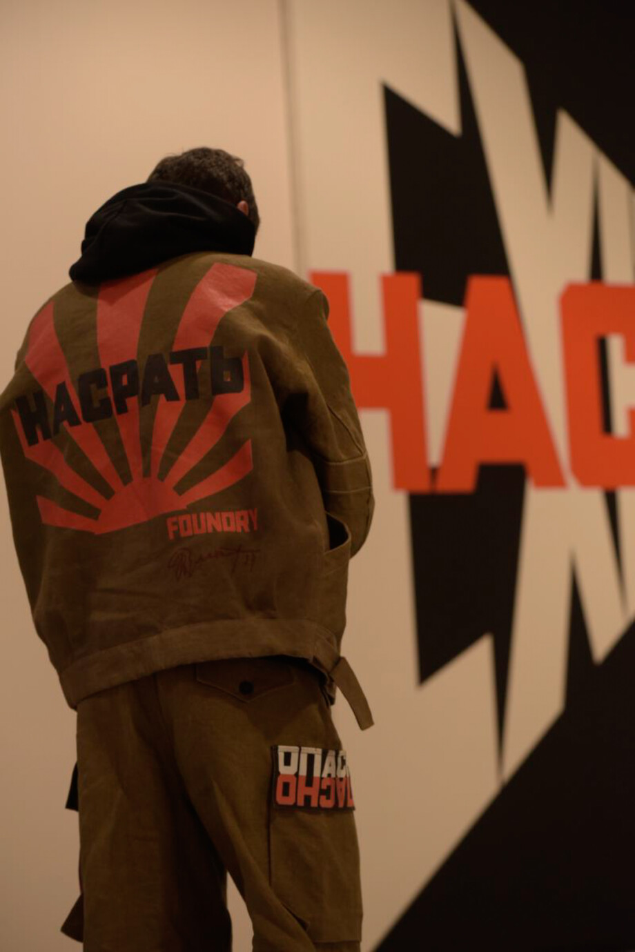 An image of someone wearing a FOUNDRY UNIFORM outfit in-front of an Erik Bulatov Painting. The outfit include a drab green overall emblazoned with artworks by Erik Bulatov, shown as part of Black Horizon BPS22